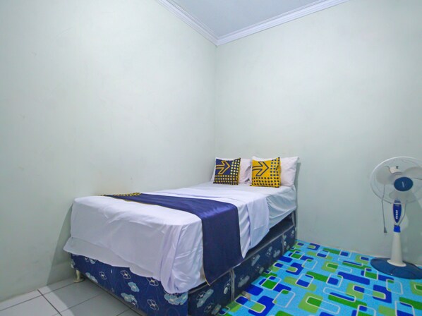 Una habitación pequeña con cama individual, colcha blanca, dos almohadas con diseño de flechas, un ventilador de pie y piso estampado.