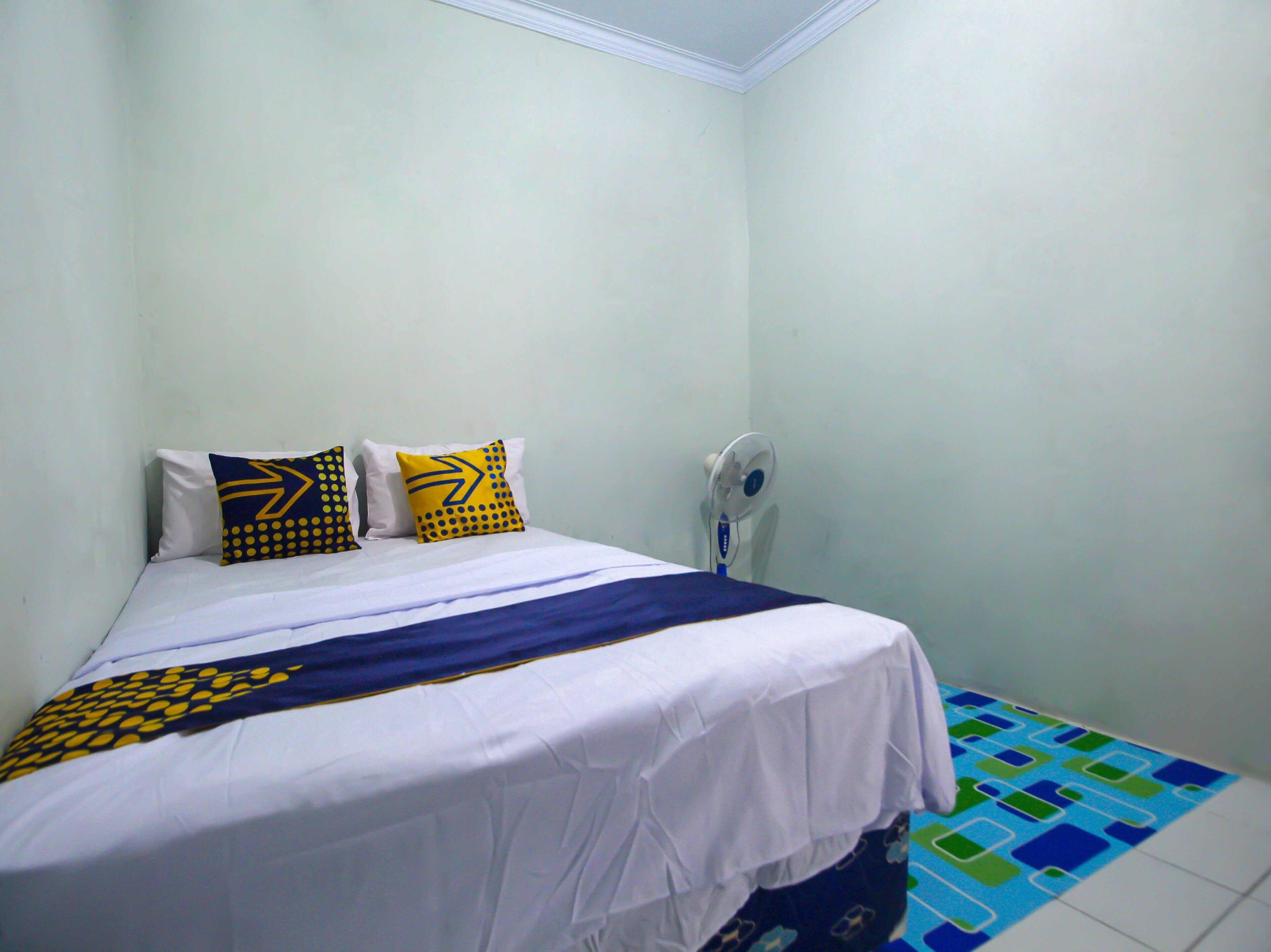 A single bed with white bedding and two decorative pillows, a standing fan, and a patterned floor.