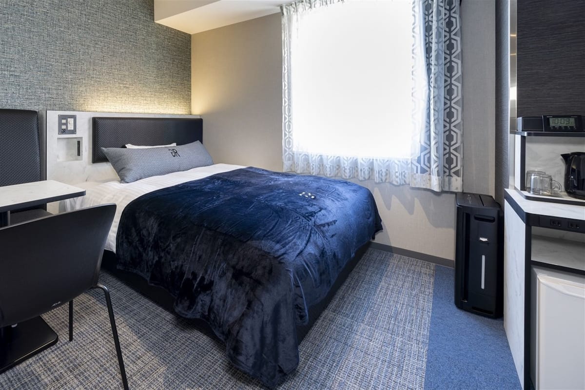Una habitación de hotel moderna con cama, escritorio, silla, kitchenette pequeña y ventana con cortinas estampadas.