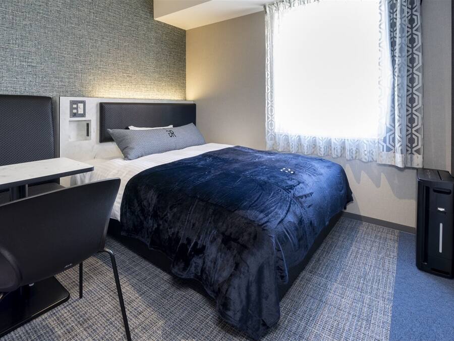 Una habitación de hotel moderna con cama, escritorio, silla, kitchenette pequeña y ventana con cortinas estampadas.