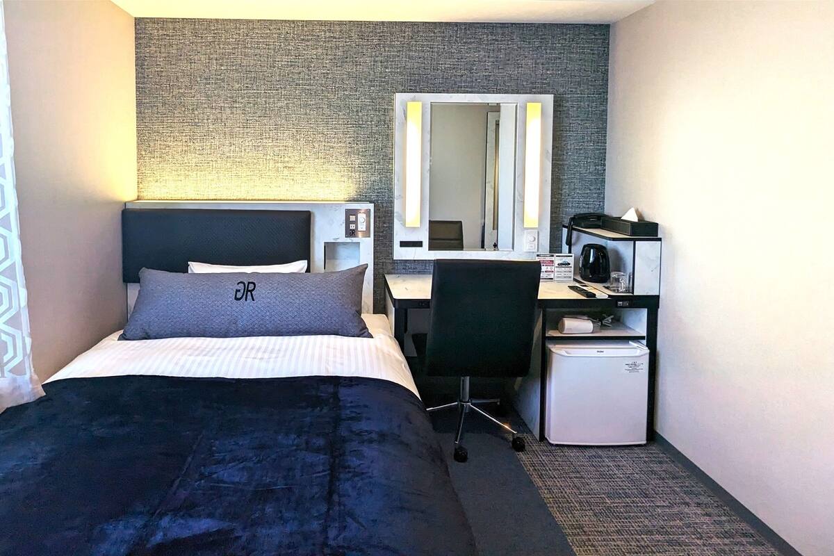 Una habitación de hotel con cama, escritorio, silla, refrigerador pequeño y espejo.