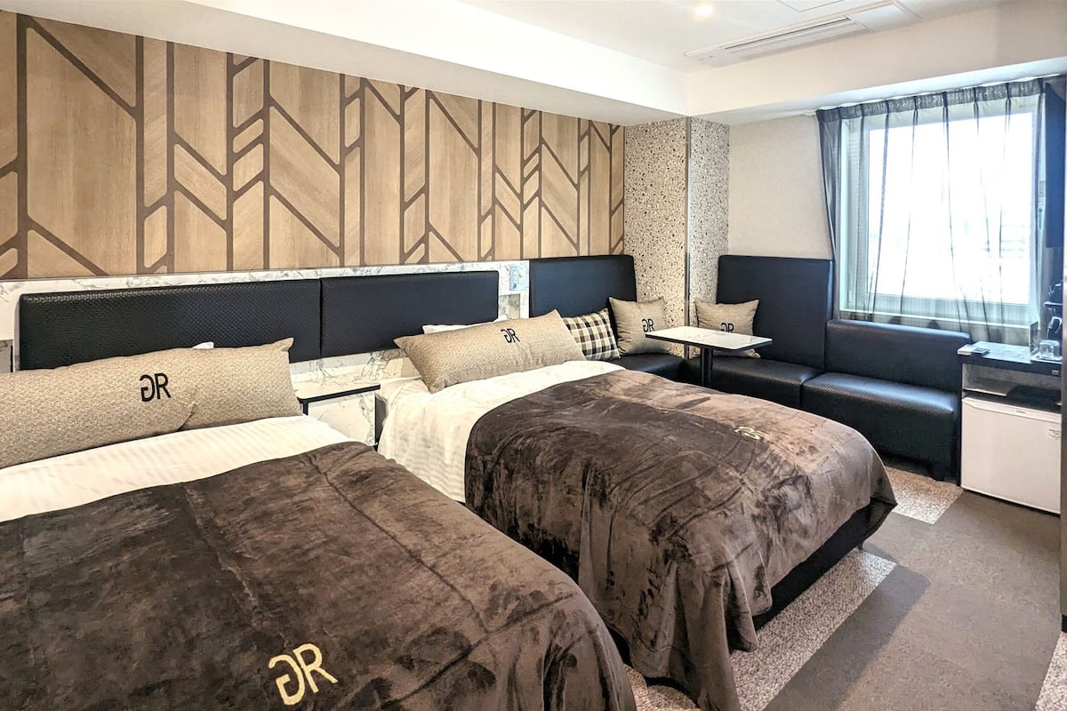 Una habitación de hotel con dos camas, una mesita, un sofá y un ventanal con cortinas.
