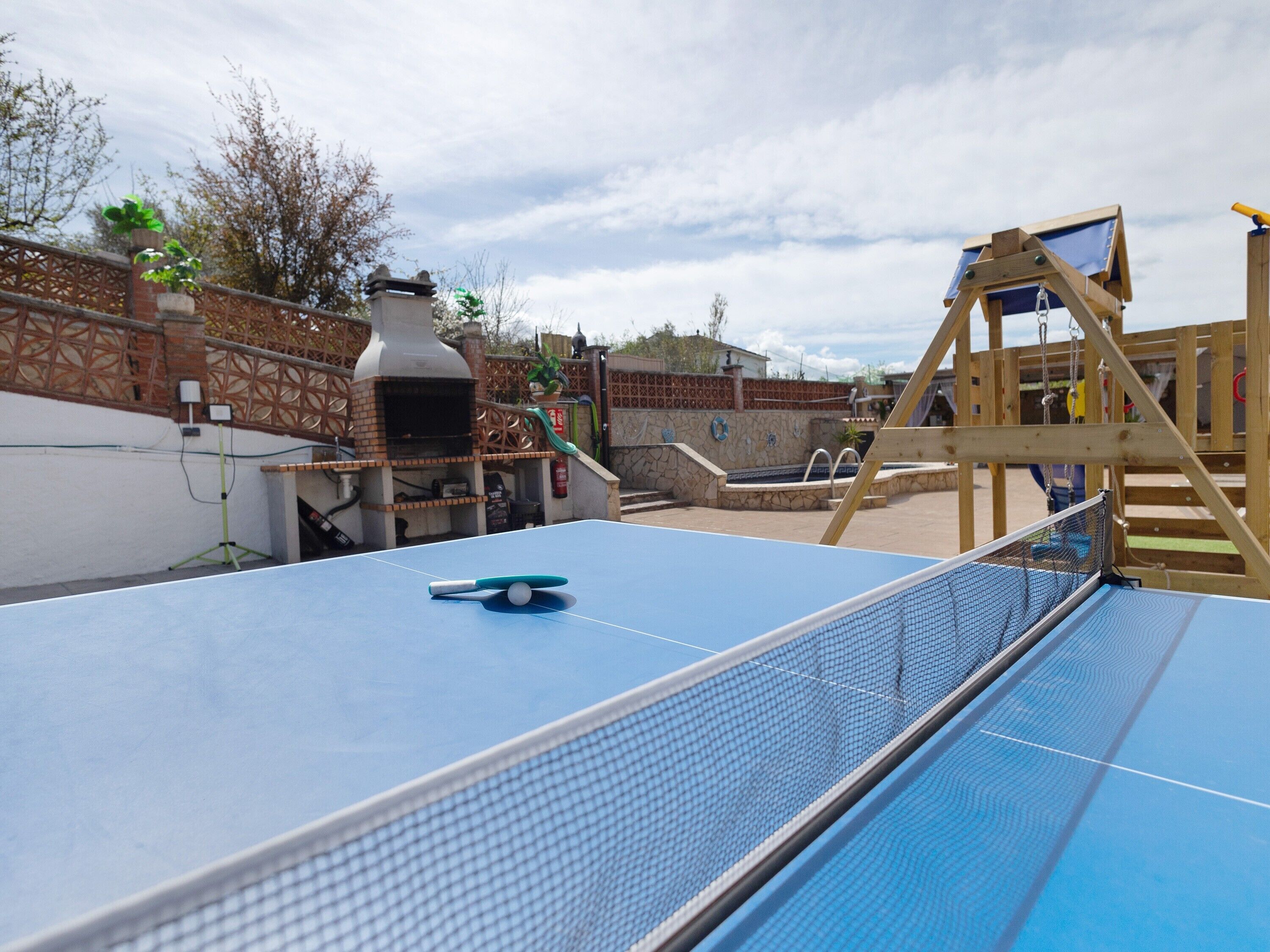 A ping pong table with a net, a wooden swing set, and a brick oven in a backyard.