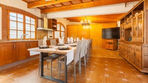 A dining area with a long table set for six, wooden cabinets, a television, and a chandelier. - Villa 'Cal Ramon for 15 Guests' with Private Pool, Wi-Fi & Air Conditioning (Rubi , Barcelona)