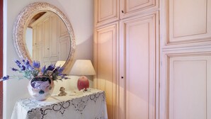 Una habitación con espejo, una mesa con mantel estampado, un florero con flores, una lámpara y un armario.
