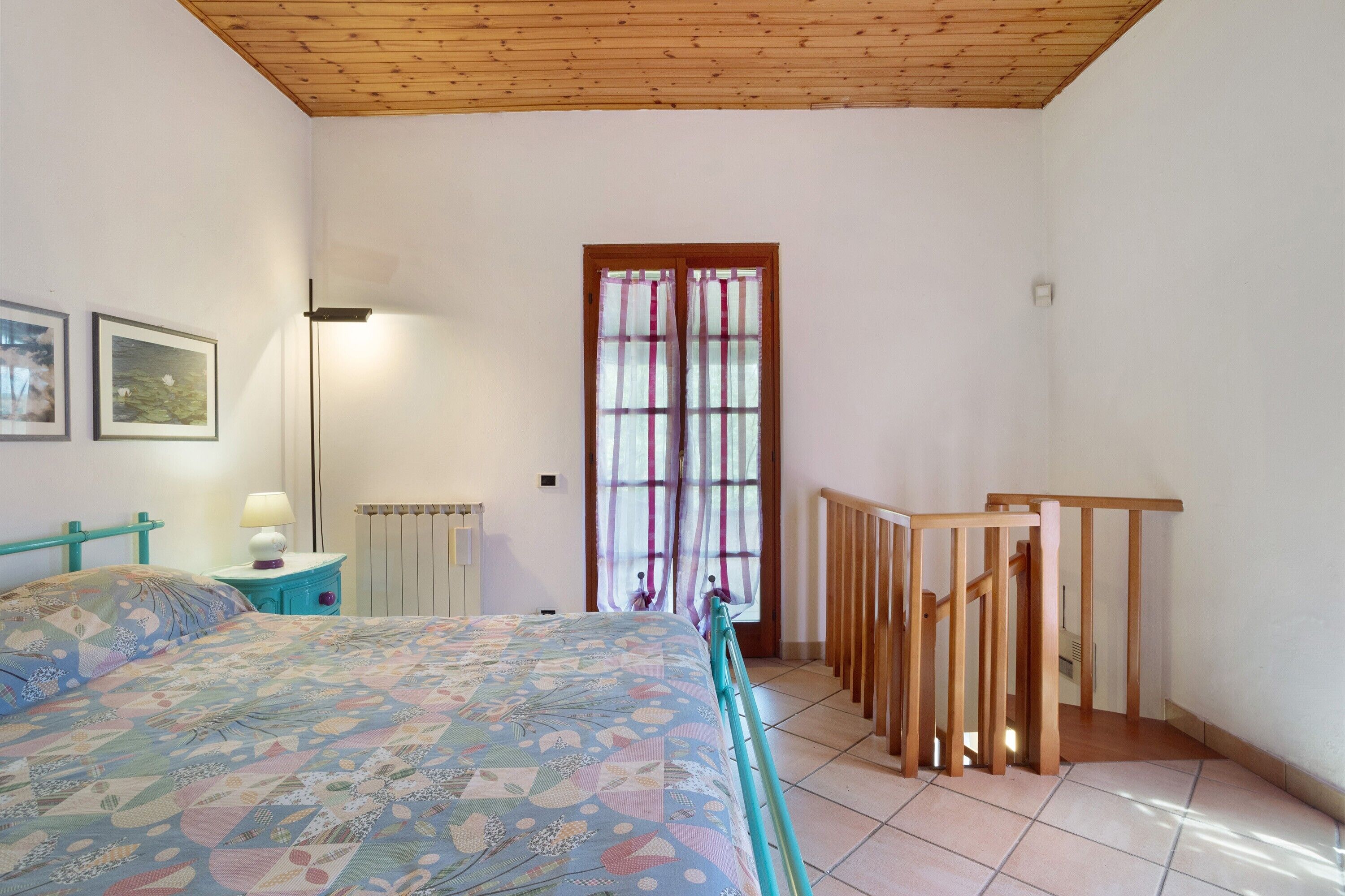 A bedroom with a bed, a nightstand, a lamp, a radiator, a window with curtains, and a wooden ceiling.