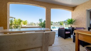 Un espace extérieur aménagé, partiellement abrité, avec vue sur la piscine, comprenant une table et des chaises, des plantes en pot et un canapé en osier.