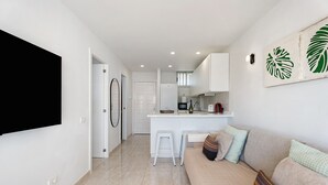 Eine moderne Küche mit weißen Schränken, einer weißen Arbeitsplatte und einem Kühlschrank. Ein weißer Hocker und ein beiges Sofa mit Kissen sind ebenfalls vorhanden.