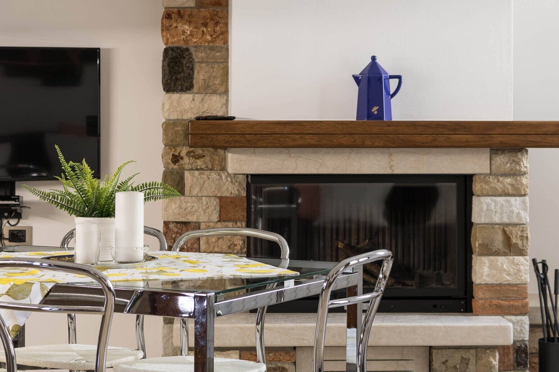 A stone fireplace with a blue kettle, a glass table with chairs, and a fern plant.