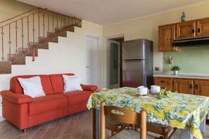 A living room with a red sofa, a dining table with a floral tablecloth, a kitchen with wooden cabinets, and a stainless steel refrigerator.