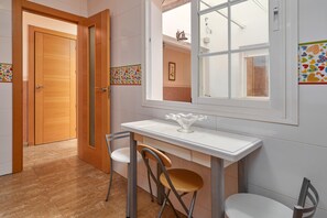 Una habitación con puerta de madera, una mesa blanca con dos sillas, una ventana con vistas y suelo de baldosas.