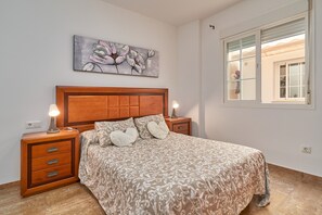 Une chambre à coucher avec une tête de lit en bois, un lit avec une literie à motifs, des lampes de chevet, une table de chevet avec des tiroirs, un tableau au mur et une fenêtre donnant sur un paysage.
