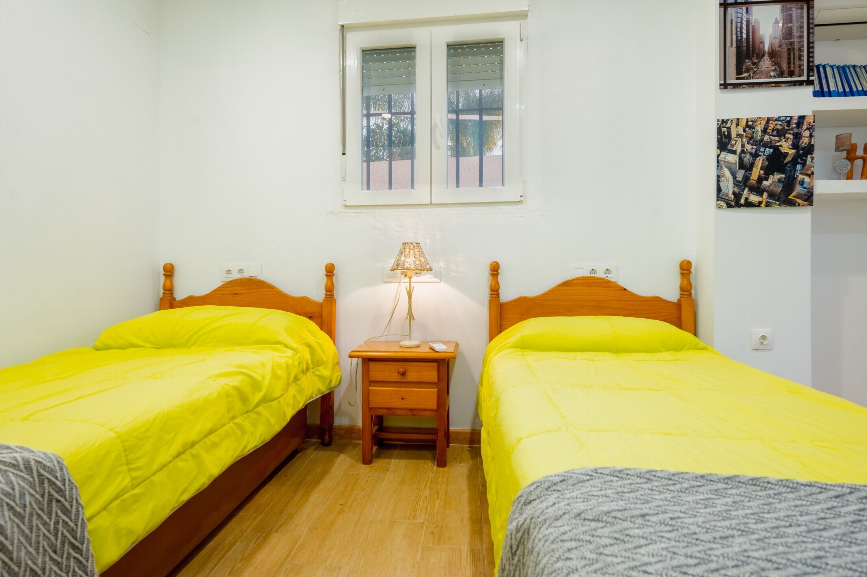 Een kamer met twee bedden, een nachtkastje met een lamp, een raam met jaloezieën en een wand met ingelijste foto's.