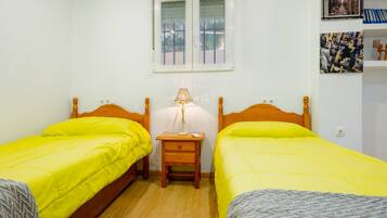 Een kamer met twee bedden, een nachtkastje met een lamp, een raam met jaloezieën en een wand met ingelijste foto's.