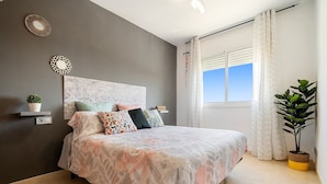 Un dormitorio con cama, cabecera, estante fijado a la pared con una planta, ventana con cortinas y planta en maceta.