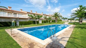 Una piscina con terraza de piedra, rodeada de césped cuidado y palmeras, con un edificio al fondo.