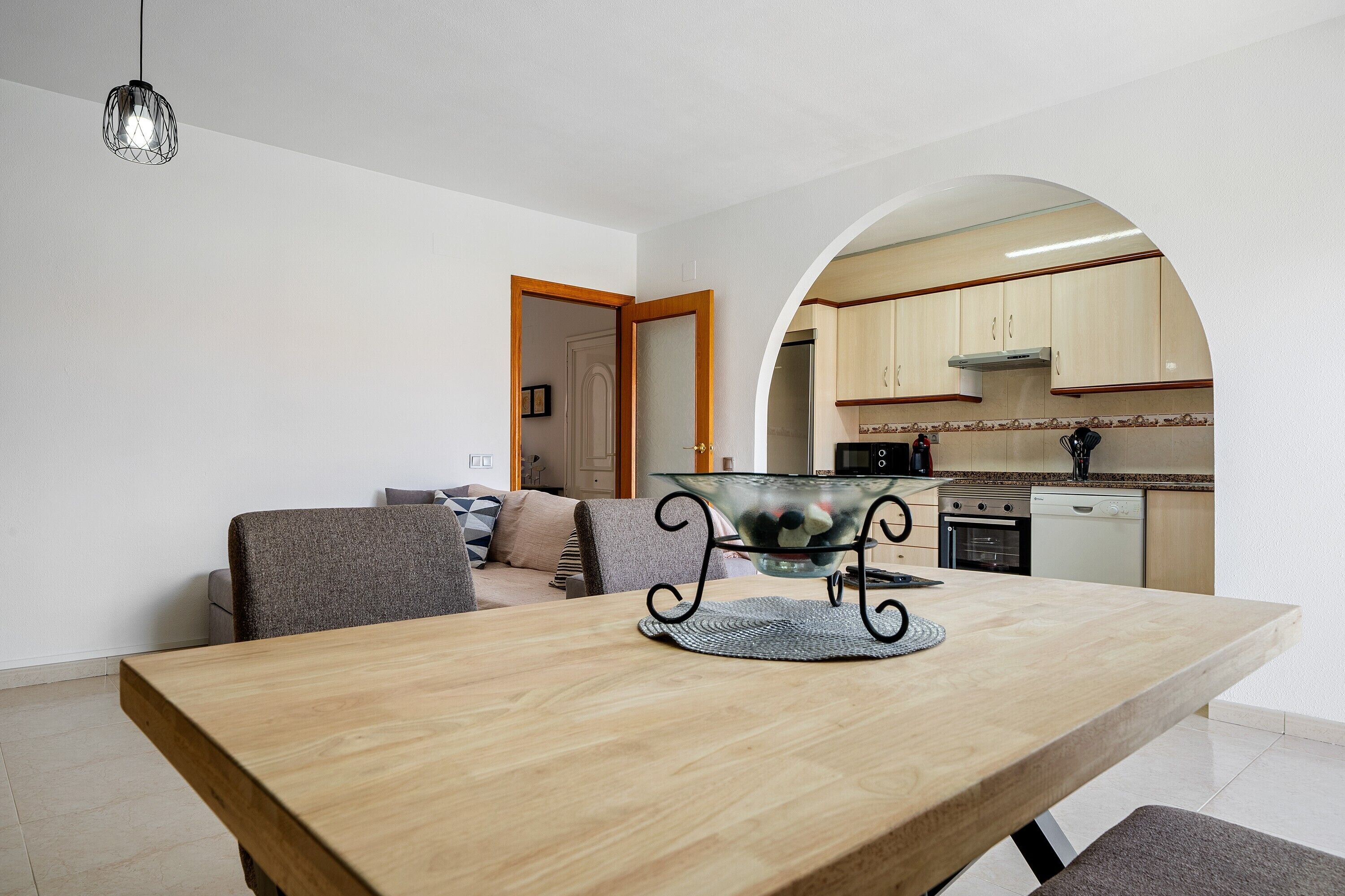 A dining area with a wooden table, a glass bowl on a metal stand, a sofa with cushions, and a kitchen visible through an archway.