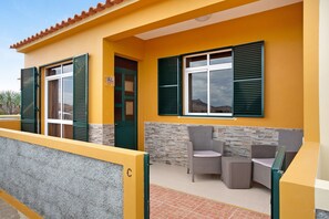 Un bâtiment jaune avec une porte, deux fenêtres avec des volets et des sièges à l’extérieur.