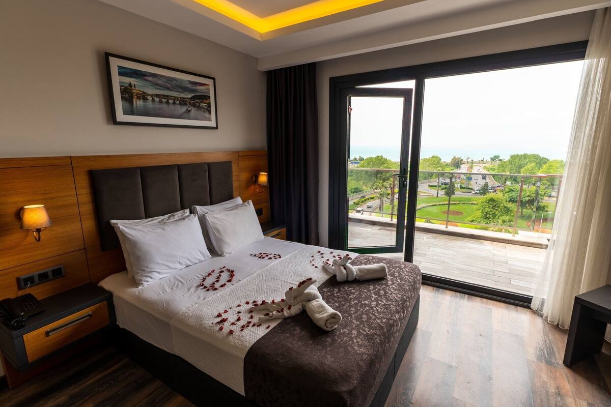 A hotel room with a large bed, a headboard, a bedside table, a lamp, and a window with a view of a cityscape.
