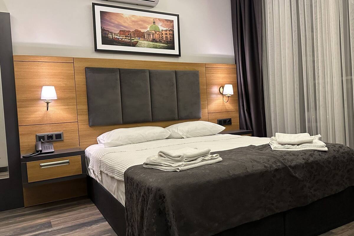 A hotel room with a large bed, two bedside lamps, a telephone, a framed picture on the wall, and a window with curtains.