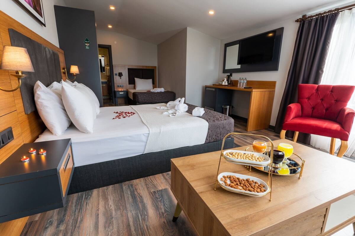 A hotel room with a bed, bedside tables, a desk with breakfast items, a red armchair, and a TV.
