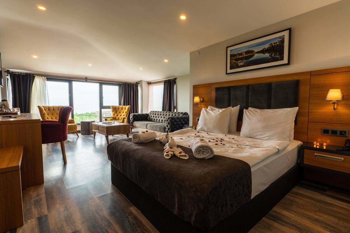 A hotel room with a large bed, a desk, a chair, and a sofa. There is a painting on the wall and a window with curtains.