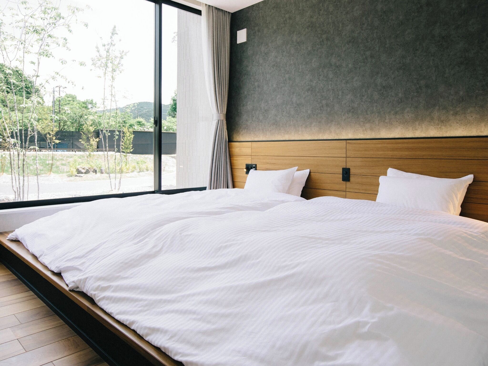 A large bed with white linens in a room with a view of greenery outside.