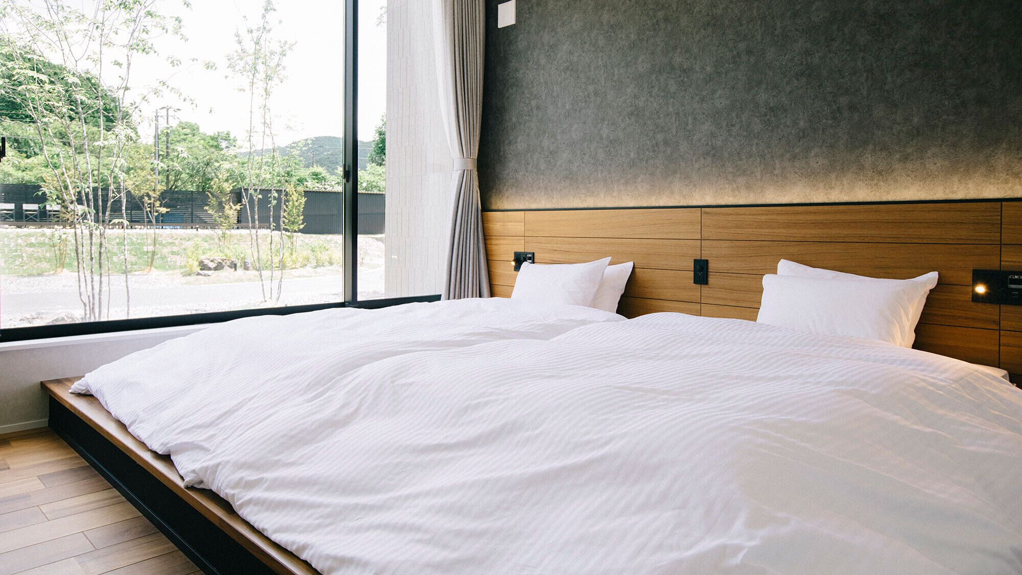 A large bed with white linens, a wooden headboard, and a view of greenery outside.