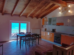 A kitchen with wooden beams, a dining area with chairs, a view through a glass door, and a kitchen counter with appliances.