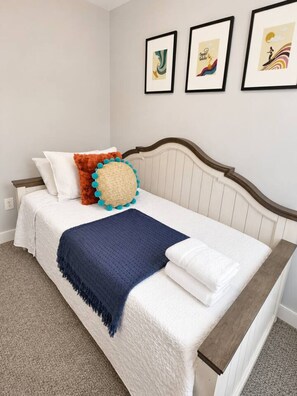 Un lit bien fait, avec une couette blanche, un plaid bleu et un coussin décoratif orné de pompons bleus.