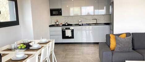 Una cocina moderna con horno integrado, microondas y lavabo. Hay una zona de comedor con una mesa preparada para dos, sillas blancas y un sofá con cojines.