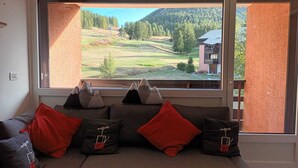 Una habitación con un ventanal amplio que ofrece vistas a un paisaje verde y un edificio. Hay un sofá con varios cojines y una alfombra.