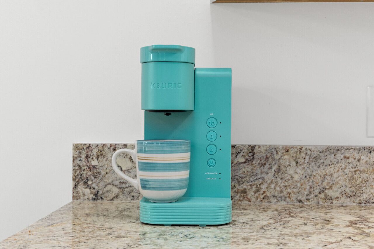Une machine à café Keurig de couleur turquoise, accompagnée d’une tasse assortie, posée sur un comptoir en granit.