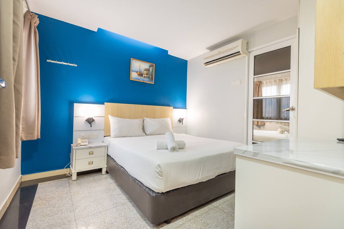 A bedroom with a large bed, bedside tables, a nightstand, a painting on the wall, and an air conditioning unit.