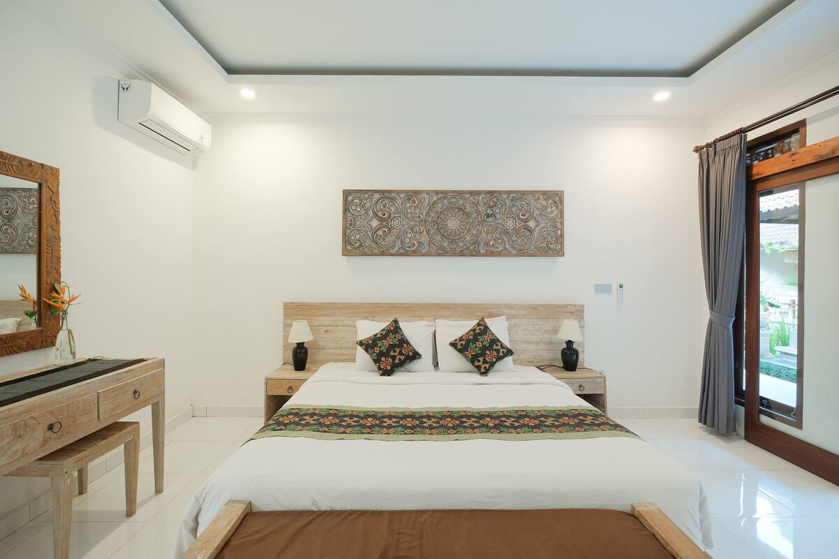 A bedroom with a wooden headboard, a large bed with floral bedding, a bedside table with a lamp, and a wall-mounted air conditioner.