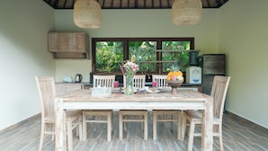 A dining area with a wooden table and chairs, a vase of flowers, and a view of the outdoors through large windows.