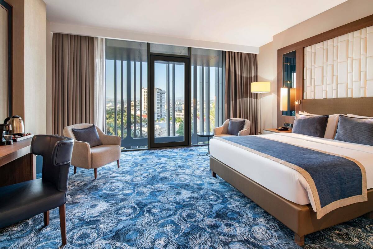 A hotel room with a large bed, a desk with a chair, a sofa, and a view of the city through large windows.