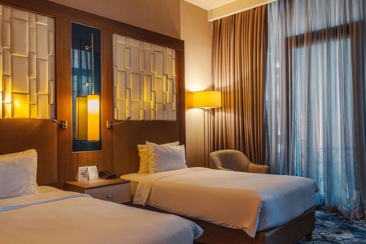 A hotel room with two beds, a desk with a phone, a chair, a lamp, and a window with curtains.