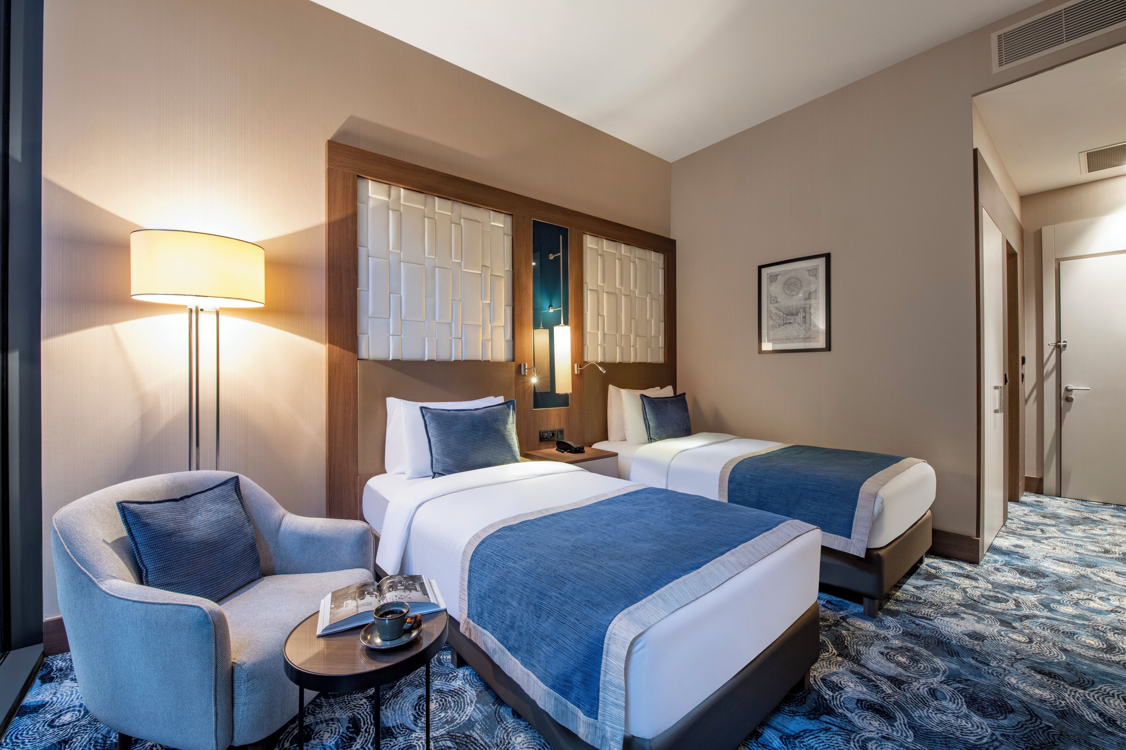 A hotel room with two beds, a sofa, a lamp, a small table, and a patterned carpet.