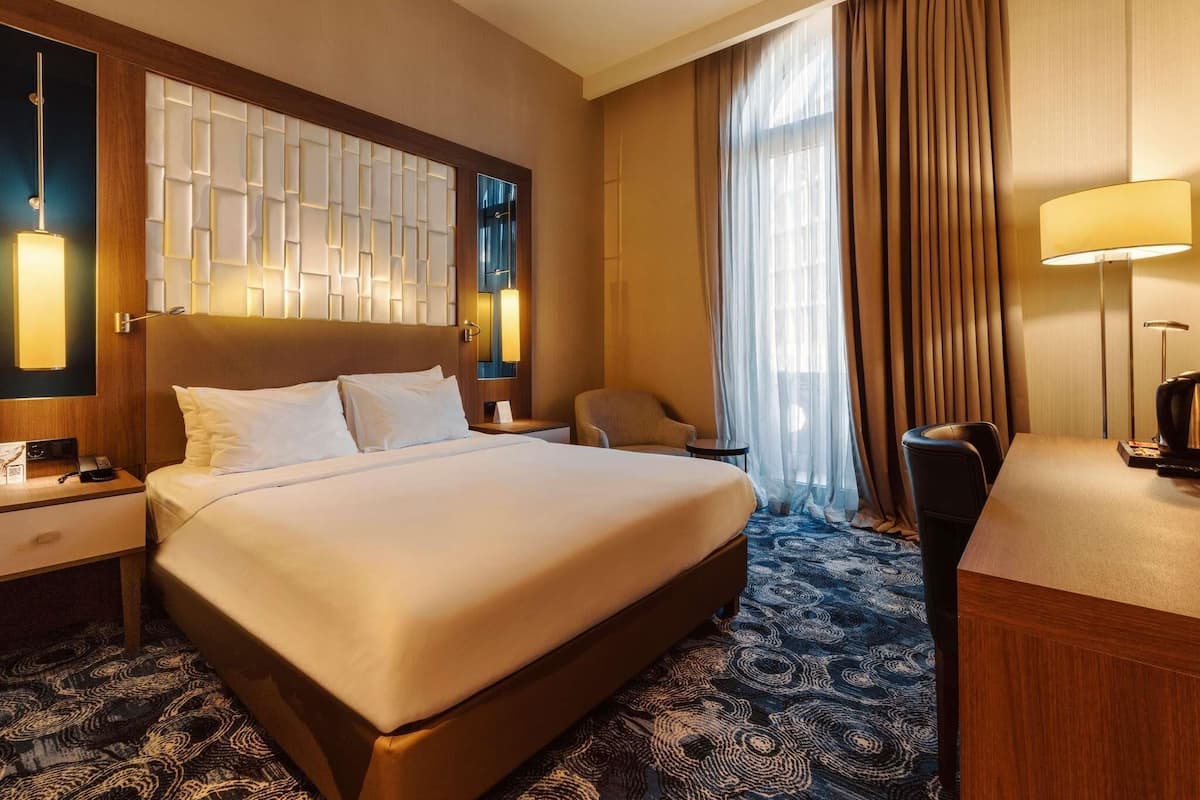 Ein Hotelzimmer mit einem großen Bett, einem Schreibtisch, einem Stuhl, einer Schreibtischlampe und einem Fenster mit Vorhängen.