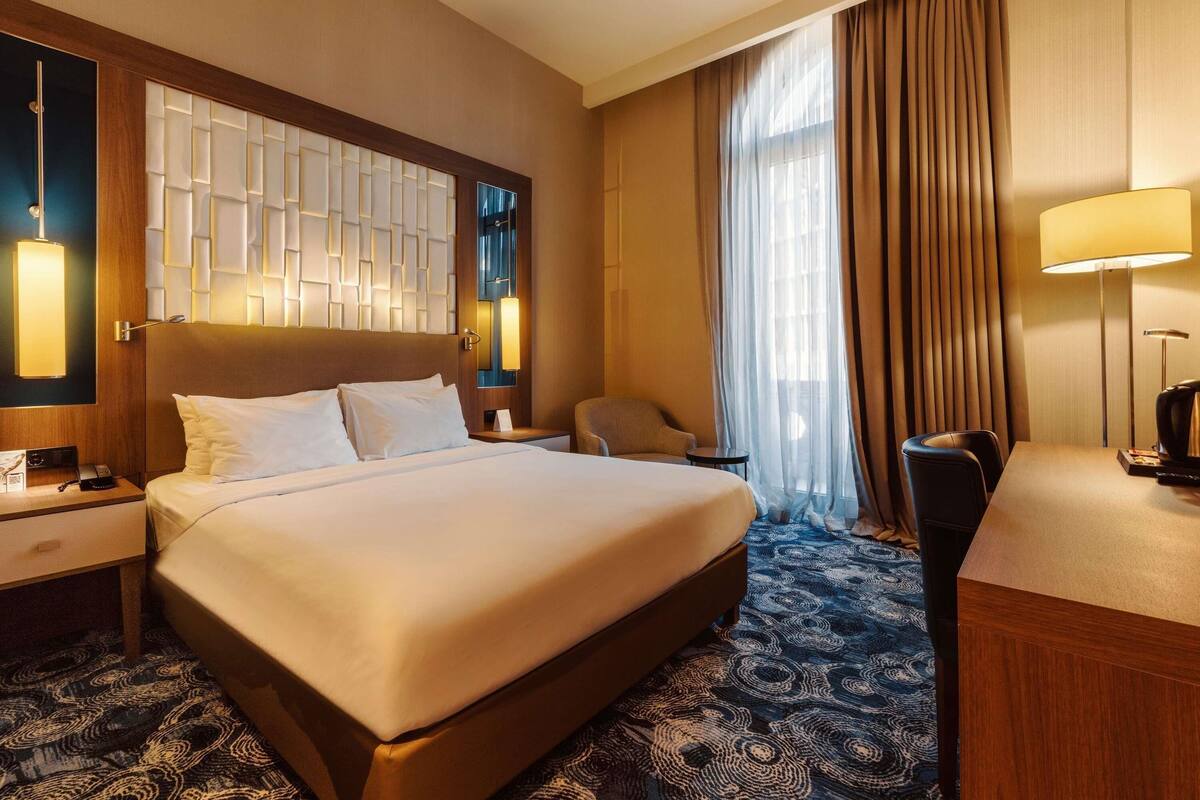 A hotel room with a large bed, a desk, a chair, a lamp, and a wall-mounted light fixture.