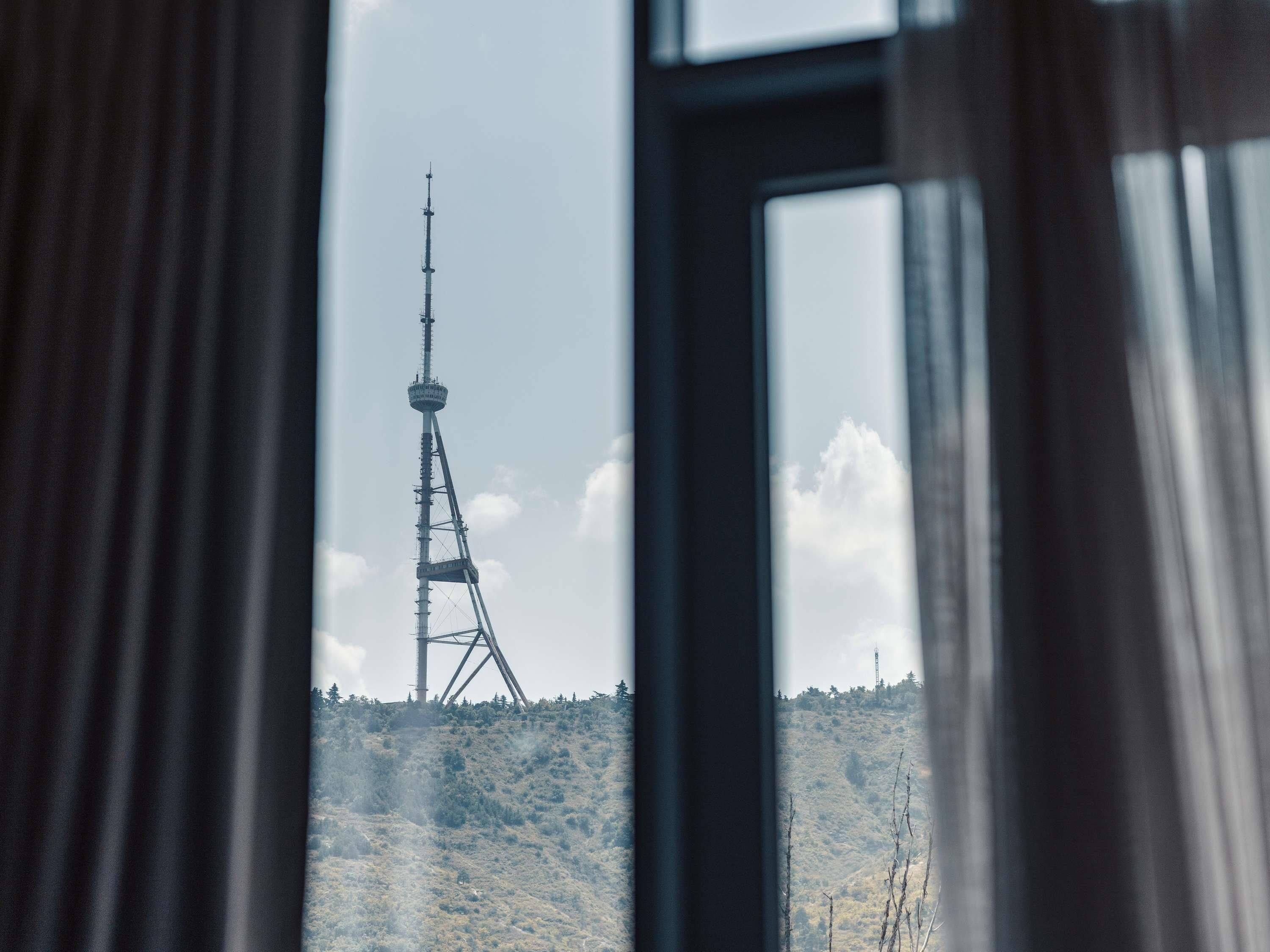 Vista a través de una ventana con cortinas, mostrando una torre de comunicaciones alta en una colina.