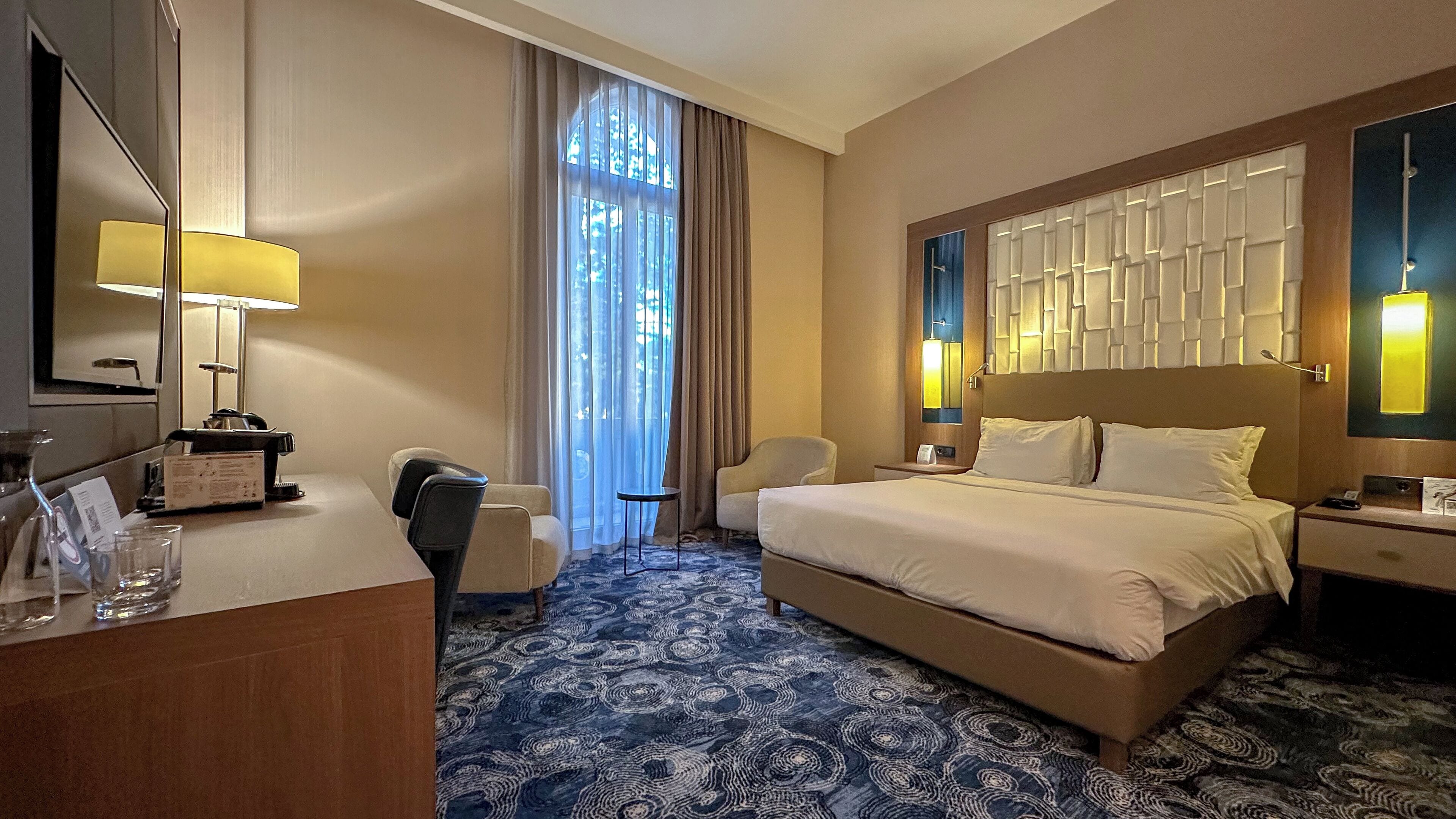 Une chambre d’hôtel avec un grand lit, un bureau avec une lampe, une chaise et une grande fenêtre avec des rideaux.