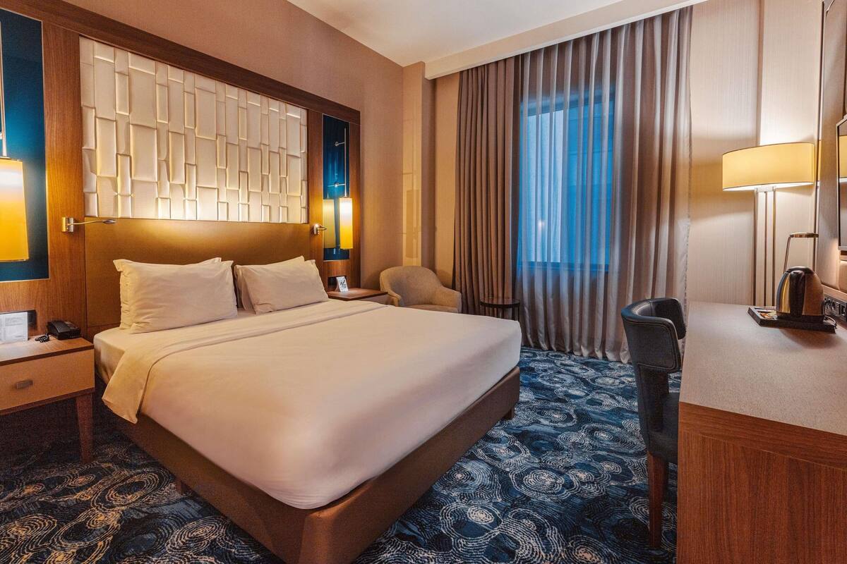 A hotel room with a large bed, a desk, a chair, a nightstand, a lamp, and a view of the outdoors.