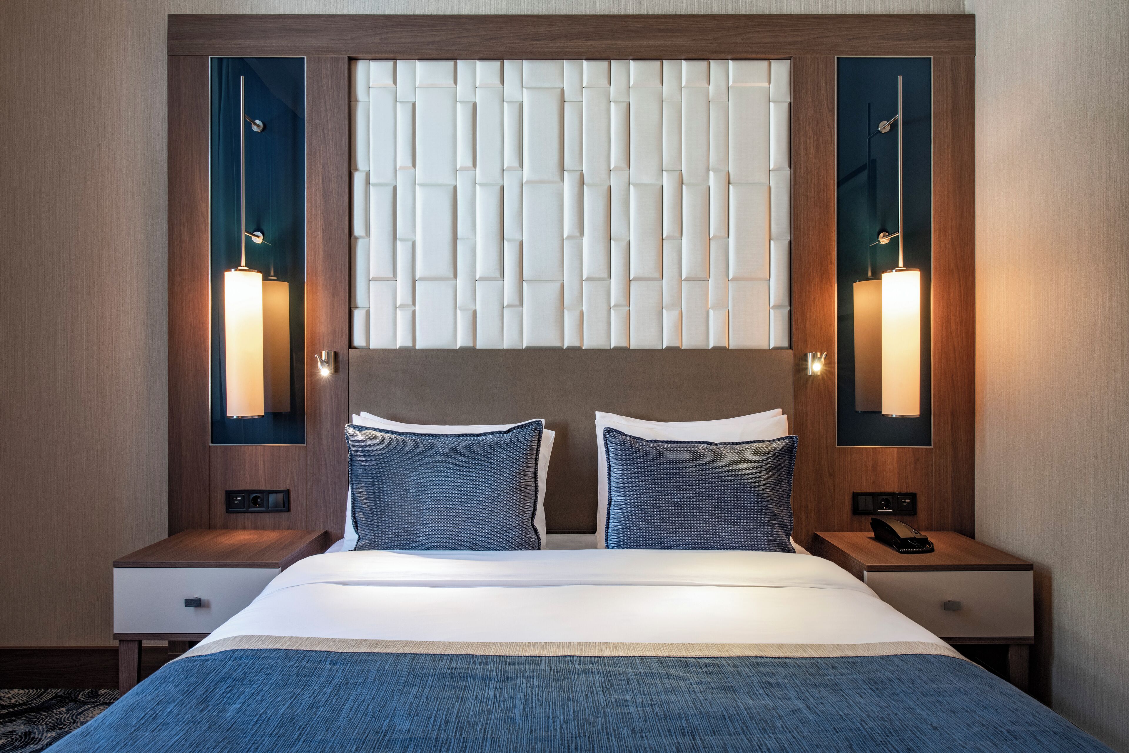 Une chambre d’hôtel avec un grand lit, deux tables de chevet, une petite table, un luminaire fixé au mur et une fenêtre avec des stores.