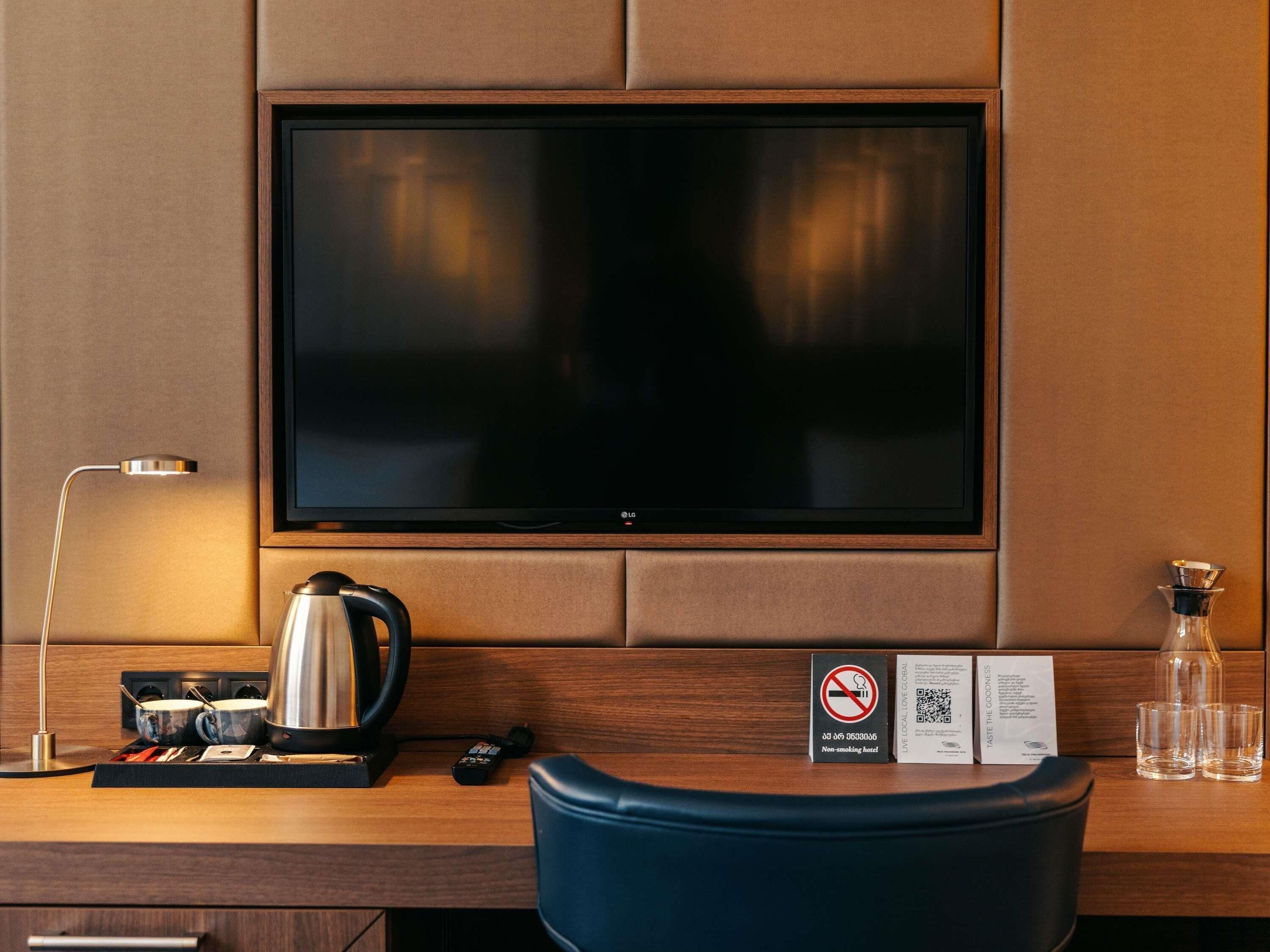 A hotel room with a flat-screen TV mounted on the wall, a coffee maker, a remote control, a non-smoking sign, and a QR code for hotel services.