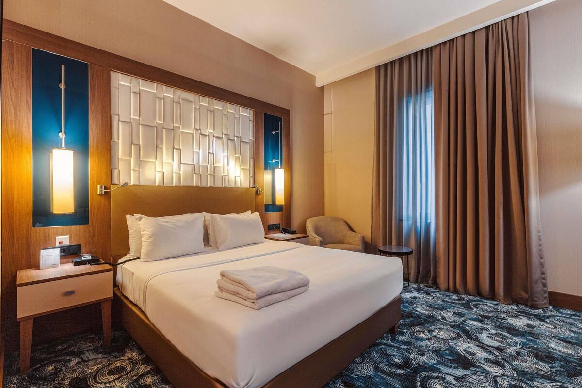 A hotel room with a large bed, a nightstand, a chair, a desk, and a window with curtains.