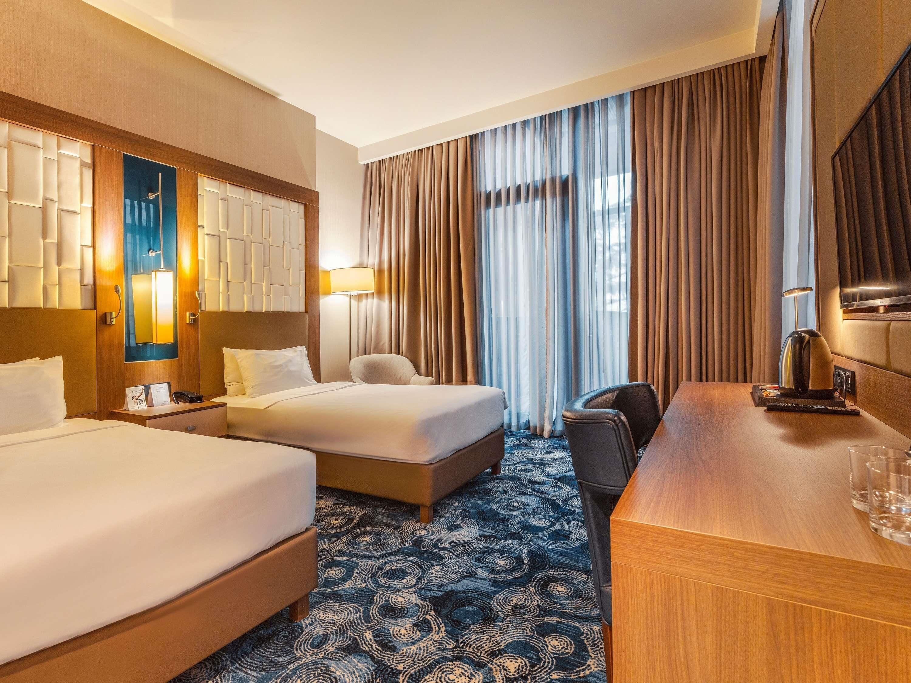 A hotel room with two beds, a desk, a chair, a TV, and a large window with curtains.