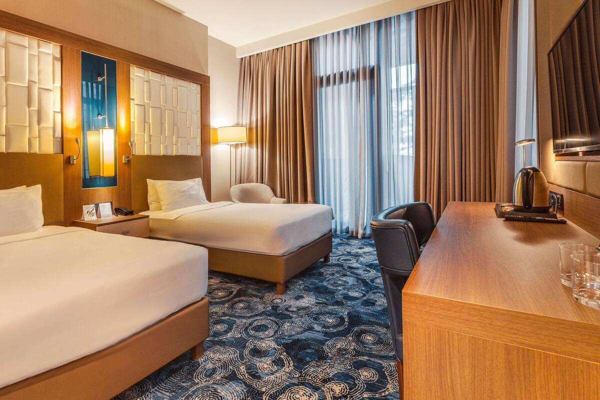 A hotel room with two beds, a desk, a chair, a TV, and a large window with curtains.