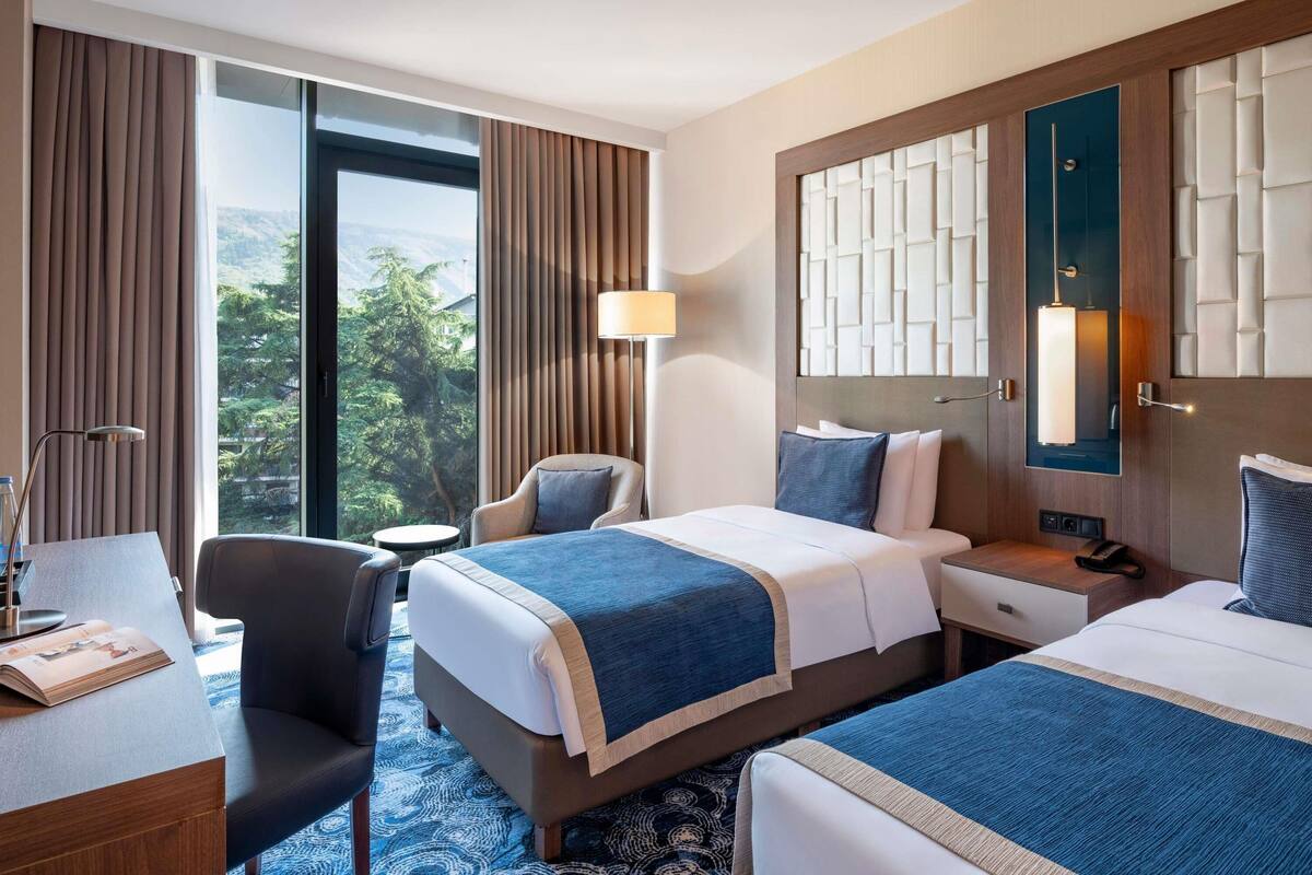 A hotel room with two beds, a desk, a chair, a lamp, and a view of greenery.